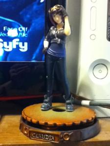 A vaguely cartoonish statue of a slender (though slightly busty) young white woman with short brown hair, dressed in blue sneakers, jeans, a blue top, and a belted, cropped dark gray jacket. She has gray gloves on both hands, and her right hand is resting on her hip, while her left hand is resting on a pair of brass goggles set on top of her head.