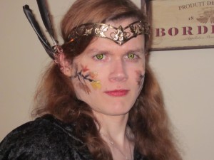 The face of a white woman with medium-length red hair, wearing a black cloak over her shoulders and a brass tiara ornamented with spirals and leaves upon her brow. Black branches and leaves in red, orange and yellow are painted on her cheeks, and she has yellow and black contact lenses in her eyes. She's wearing pointed ears made of copper wire and ornamented with black feathers.